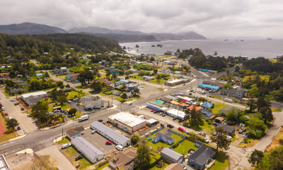 Aerial view of a coastal town with homes and other small buildings along a well-organized grid of roads. Surrounding the town are thick, green forests, rolling hills, and, in the distance, large headlands jutting out to sea. 
