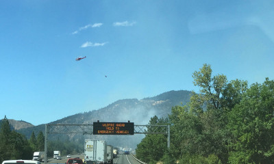 A backed-up freeway surrounded by lush vegetation on both sides, with hills partially obscured by smoke ahead. There is also a helicopter in the air and a signboard that reads, "Wildfire ahead. Yield to emergency vehicles."