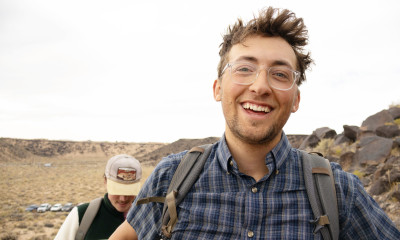 A smiling young man with glasses, a blue plaid shirt, and a backpack in an arid, rocky landscape.