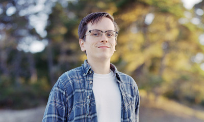 A man with black glasses, a blue flannel shirt, and a white t-shirt underneath, standing in an outdoor setting.
